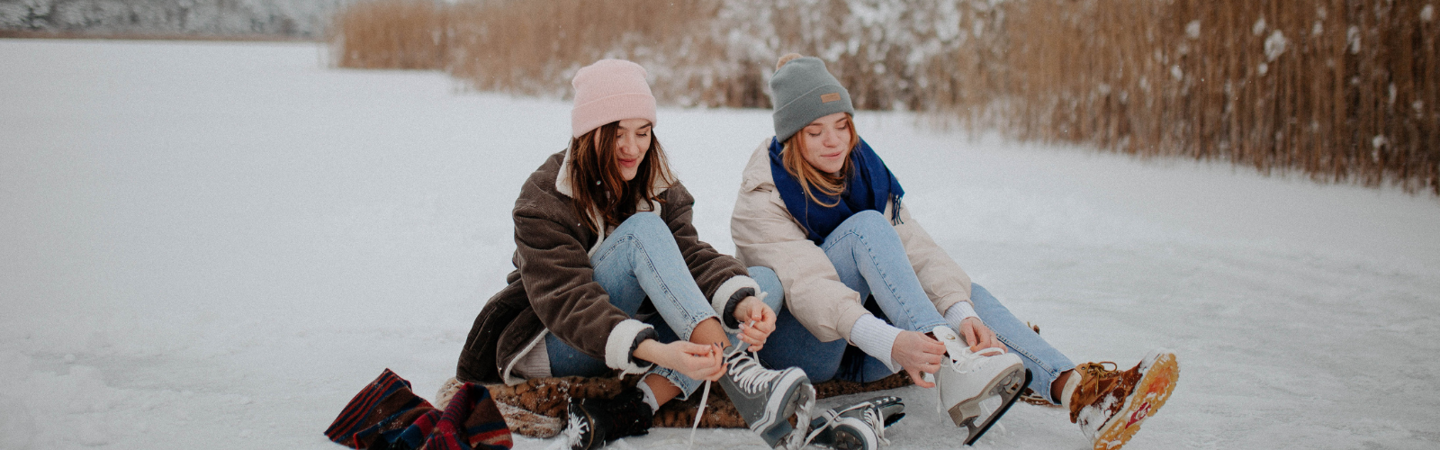 Two women sit side by side, putting on their ice skates.